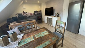 A loft-style living space with a sloped ceiling, a dining table with a rustic design, a television mounted on the wall, and a modern refrigerator. - Beautiful vacation apartment at the pulse of the Green Hell (Kelberg)