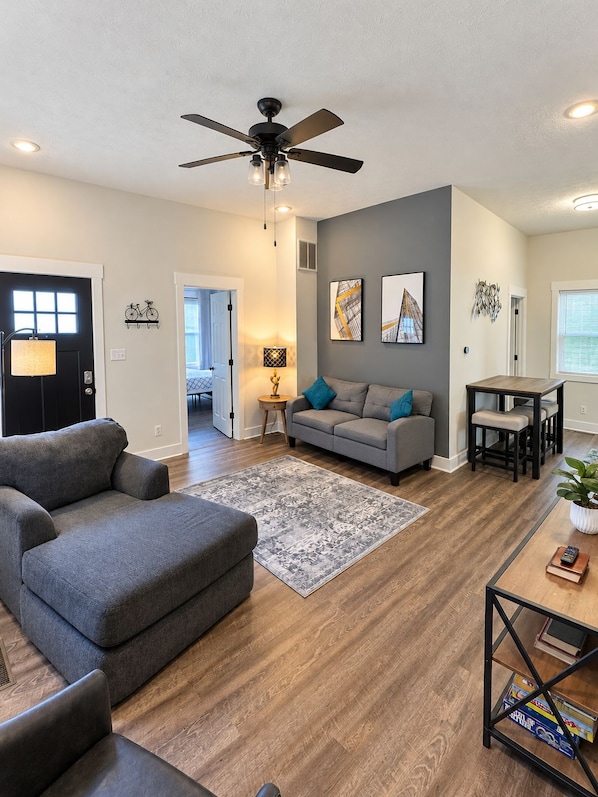 A modern living room with a grey sofa, a dining table, and a ceiling fan. - Walk to Lucas Oil Stadium - Clean 4BR Home (Indianapolis)