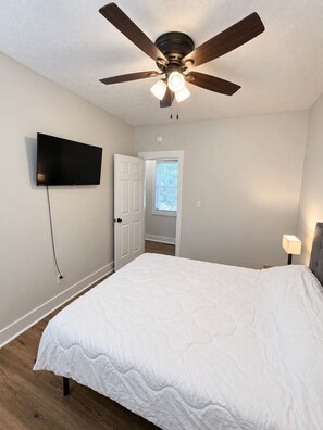 A bedroom with a bed, a ceiling fan, a TV mounted on the wall, and a door leading to another room. - Walk to Lucas Oil Stadium - Clean 4BR Home (Indianapolis)
