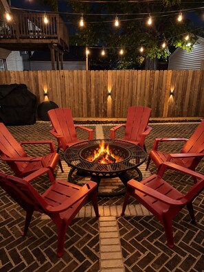 A fire pit surrounded by red chairs on a brick patio with a wooden fence and string lights in the background. - Walk to Lucas Oil Stadium - Clean 4BR Home (Indianapolis)
