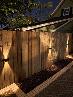 A wooden fence with integrated lighting along the base, a brick pathway, and a house in the background. - Walk to Lucas Oil Stadium - Clean 4BR Home (Indianapolis)