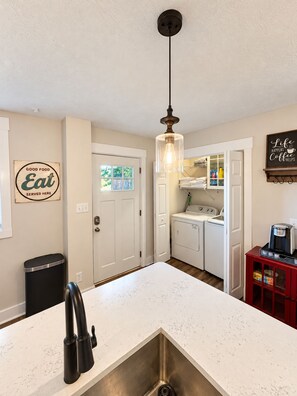 A kitchen with a stainless steel sink, a hanging light fixture, a laundry area with a washing machine, and a coffee maker on a red cabinet. - Walk to Lucas Oil Stadium - Clean 4BR Home (Indianapolis)