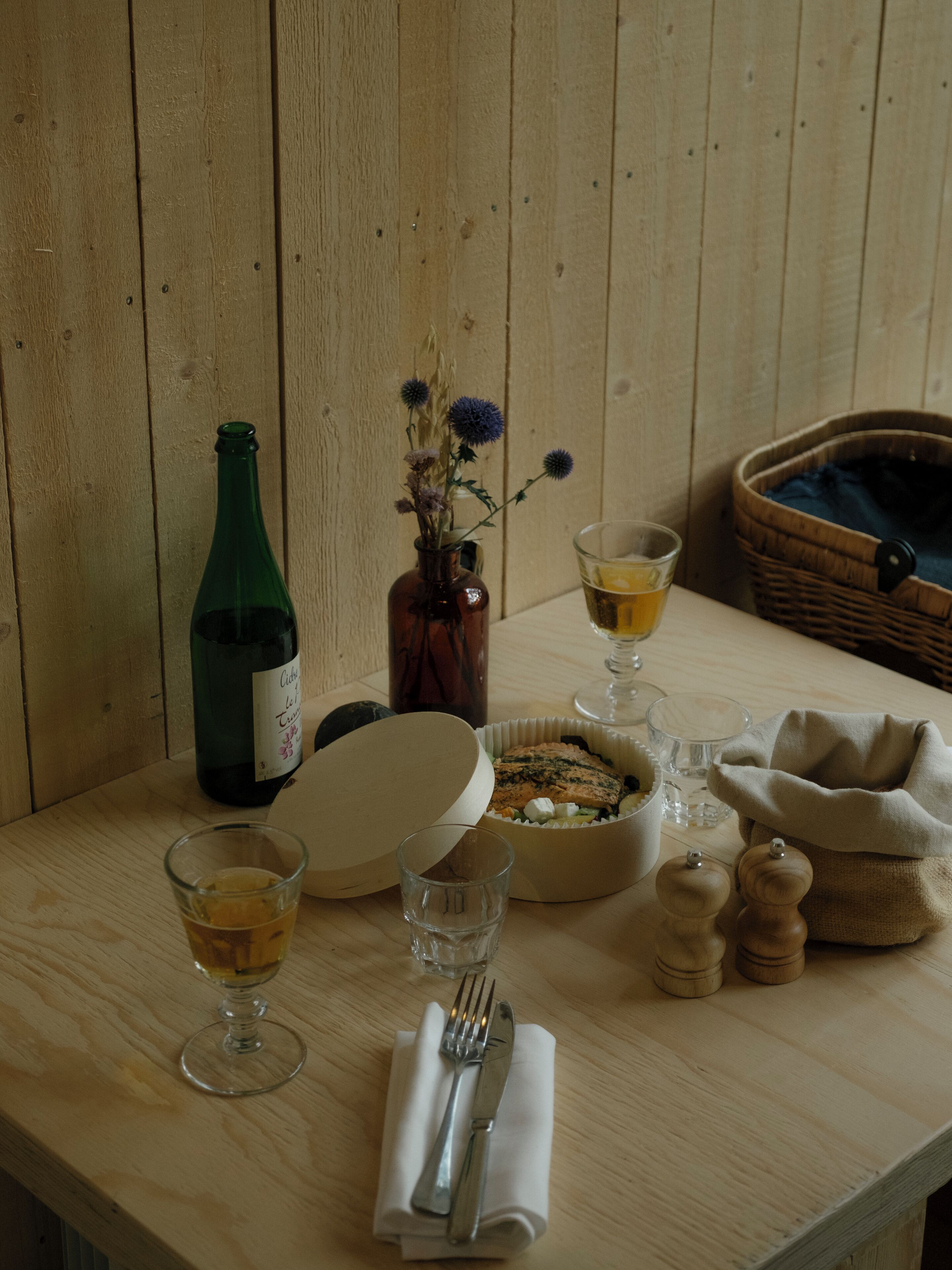A wooden table with a bottle, a bowl of food, glasses, a napkin with cutlery, and a basket.