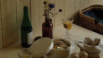 A wooden table with a bottle, a bowl of food, glasses, a napkin with cutlery, and a basket.