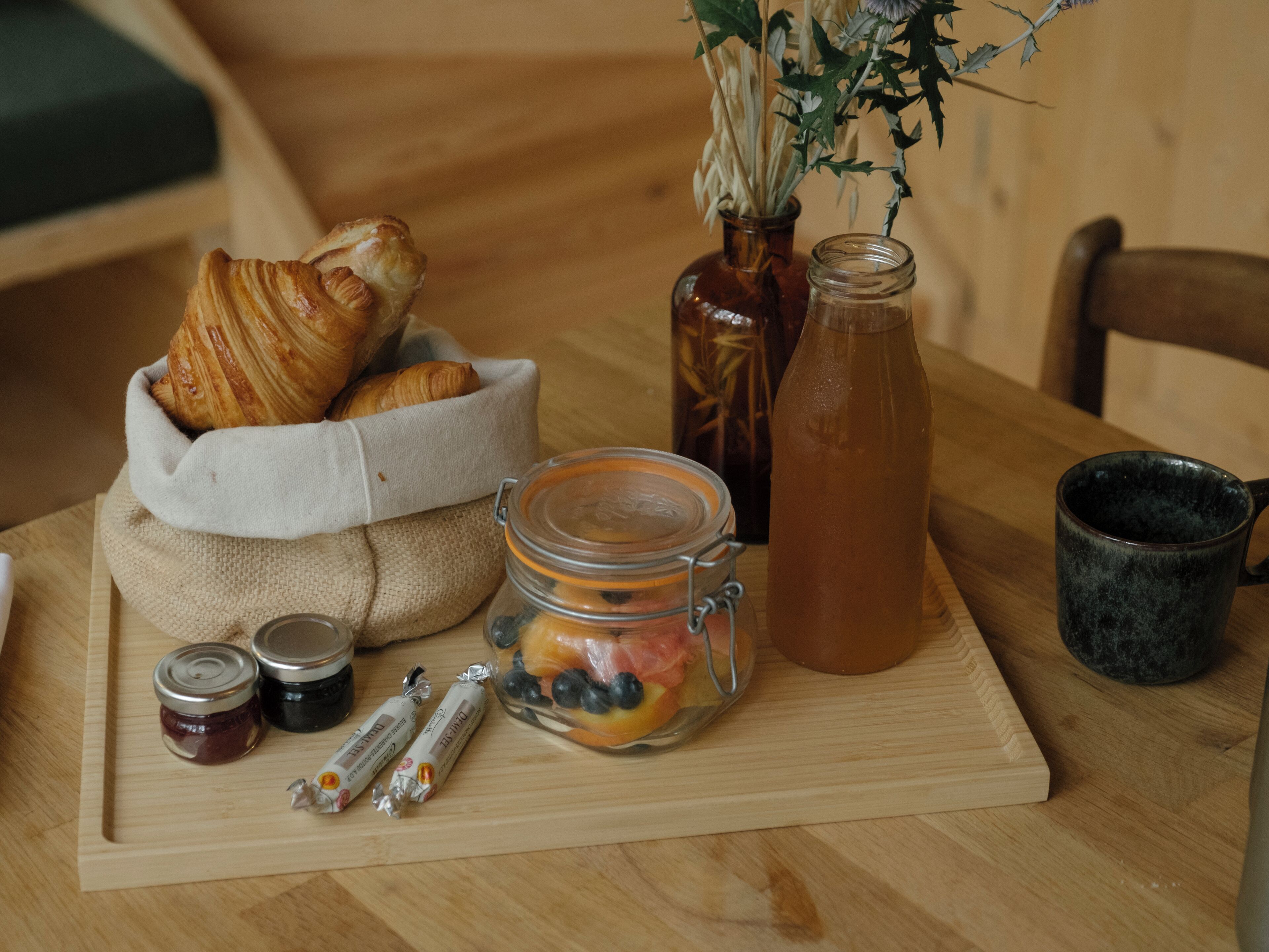 A wooden table with a basket of croissants, a glass jar of fruit, a bottle of tea, and two small jars of jam.