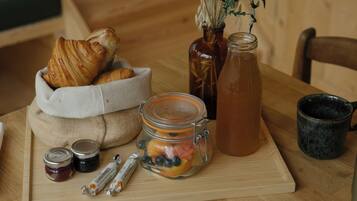 A wooden table with a basket of croissants, a glass jar of fruit, a bottle of tea, and two small jars of jam.