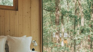 Makuuhuone, jossa on suuri sänky, puuseinät, ikkuna, josta avautuu näkymä metsään, ja yöpöytälamppu.