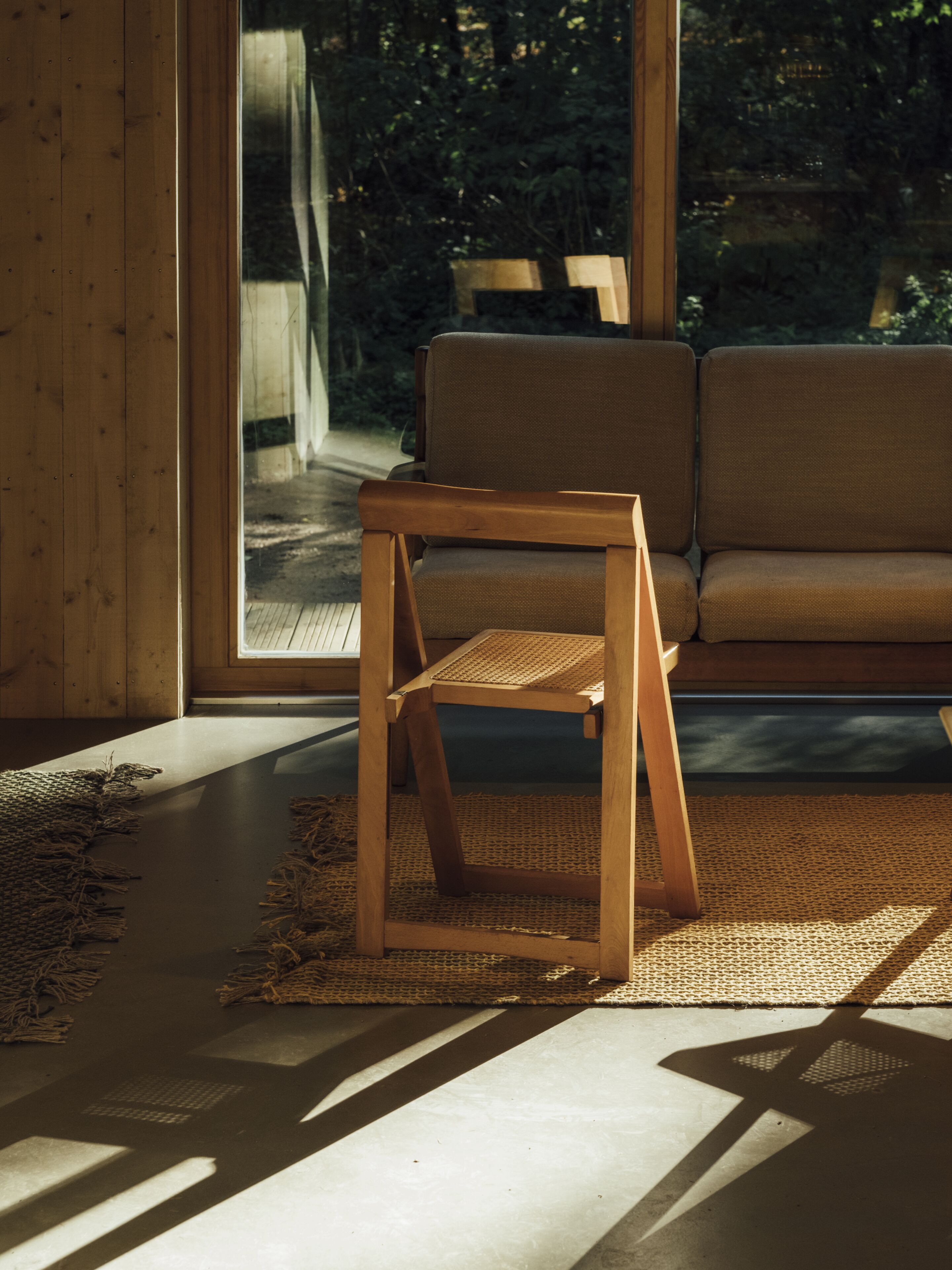 Ein Holzhocker mit gewebter Sitzfläche und Lehne, platziert auf einer Gewebematte in einem Raum mit großem Fenster, einer Couch und einem Betonboden.