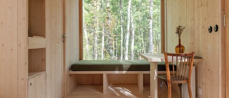A wooden cabin interior with a bench, a chair, and a window overlooking a forest.