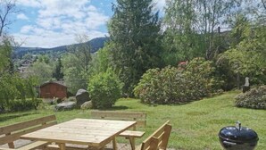 A wooden picnic table and chairs set on a stone patio with a barbecue grill. - Maison Familiale "les 3 Roches" (Xonrupt-Longemer)