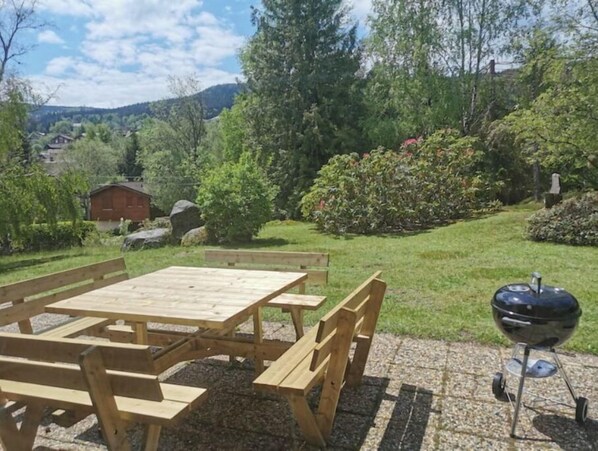 A wooden picnic table and chairs set on a stone patio with a barbecue grill. - Maison Familiale "les 3 Roches" (Xonrupt-Longemer)