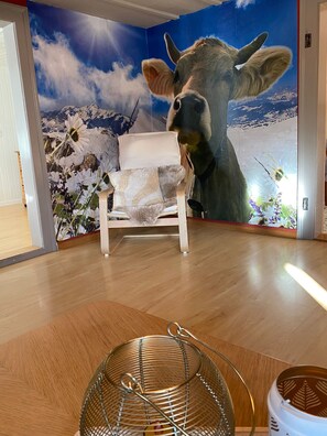 Ein Zimmer mit einer Kuh-Mural, einem weißen Stuhl und einem gewebten Korb auf Holzdielen.
