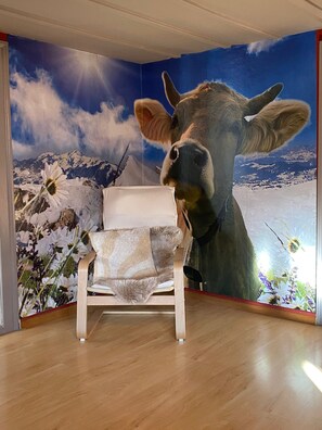 A room with a wooden floor, a white chair, and a large wall mural of a cow and mountains.