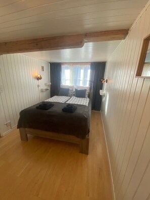 Ein Schlafzimmer mit Holzboden, einem Bett mit gemusterten Betttextilien, zwei Nachttischlampen und einem Fenster mit Vorhängen.