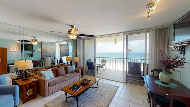 A spacious living room with a sofa, coffee table, and a view of the ocean through large sliding glass doors. - Beachfront Living at the Apollo I On-Site Sunset Grille Tiki/Restaurant (Marco Island)