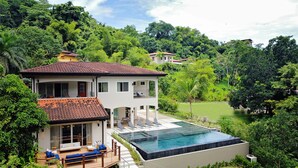 Une maison à deux étages avec une piscine, entourée d’une végétation luxuriante et de collines.