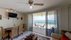 A spacious living room with a balcony overlooking the sea, furnished with wicker chairs and a dining table.