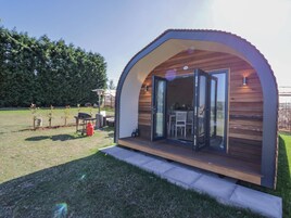 A modern, semi-open wooden structure with a glass door, a patio area with outdoor furniture, and a clear view of the surrounding greenery.