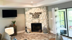 A living room with a stone fireplace, a flat-screen TV mounted on the wall, and a large window with curtains. - Modern Vibe, 5 Beds, 6 pools, Great Location (Lake Ozark)