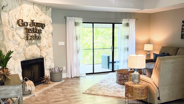 A living room with a stone fireplace, a sofa, wicker side tables, and a view through a glass door to an outdoor area. - Modern Vibe, 5 Beds, 6 pools, Great Location (Lake Ozark)