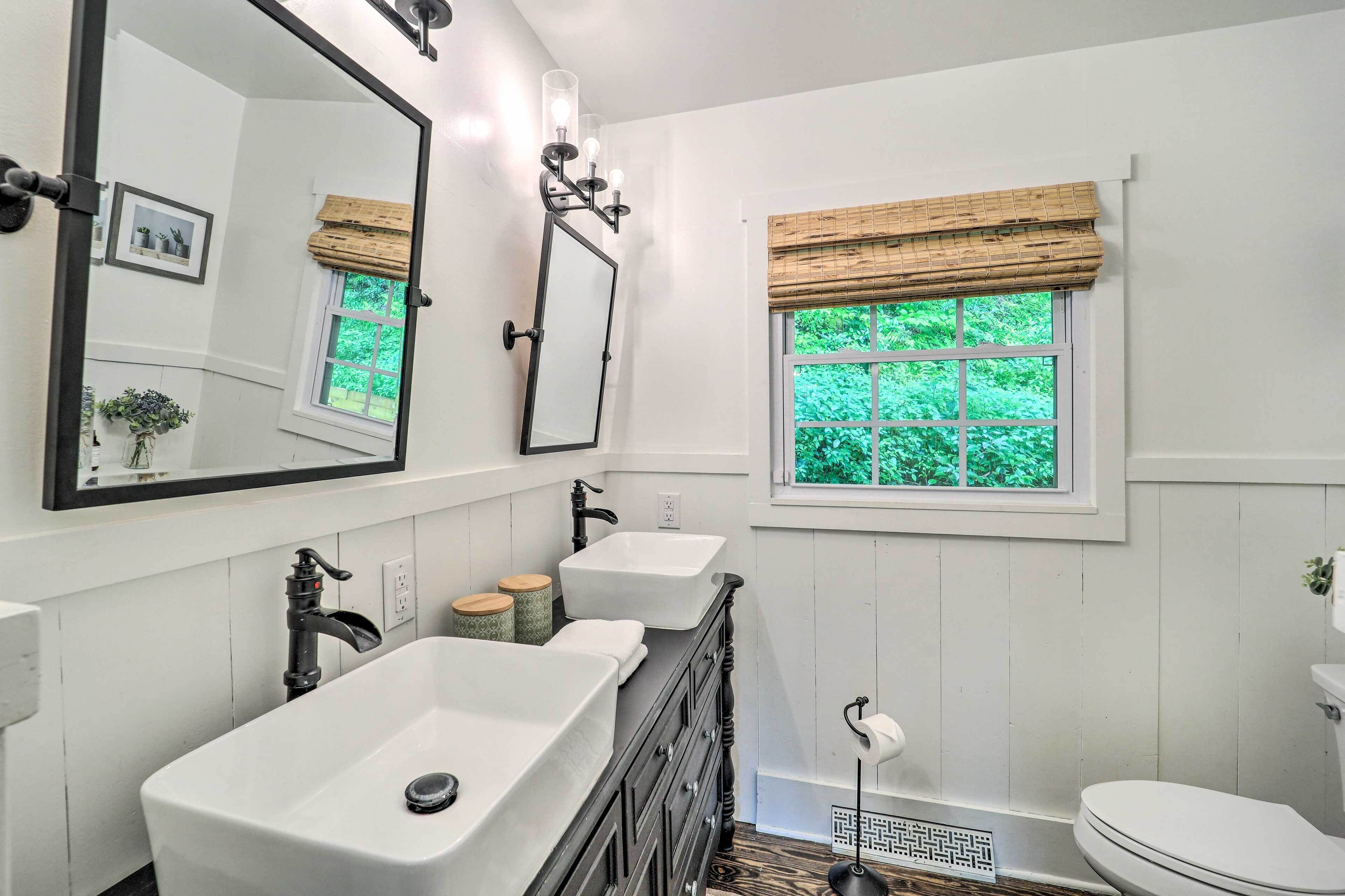 A bathroom with a large mirror, two sinks, a toilet, and a window with a view of greenery.