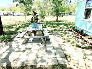 A picnic table with a flower pot on it, surrounded by a grassy yard with trees and a house in the background. - Cozy Bungalow, close to the lake. (Graford)