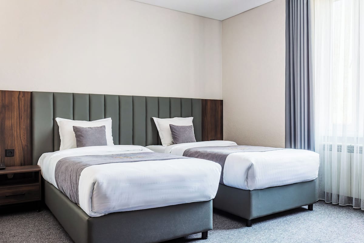 Habitación de hotel con dos camas, cabecero de madera, mesita de noche y ventana con cortinas.
