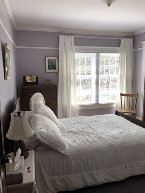 A bedroom with a bed, a dresser, a chair, a lamp, a window with curtains, and a framed picture on the wall.