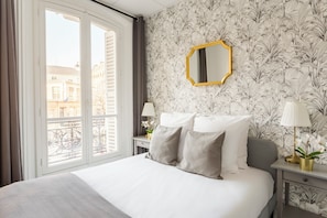 A hotel room with a bed, a nightstand with a lamp, a mirror, and a view of buildings outside. - Family Suite - Marais - Turbigo I (Paris)