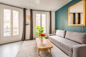 A room with a grey sofa, a wooden coffee table with a vase of flowers, and a large mirror on the wall. - Family Suite - Marais - Turbigo I (Paris)
