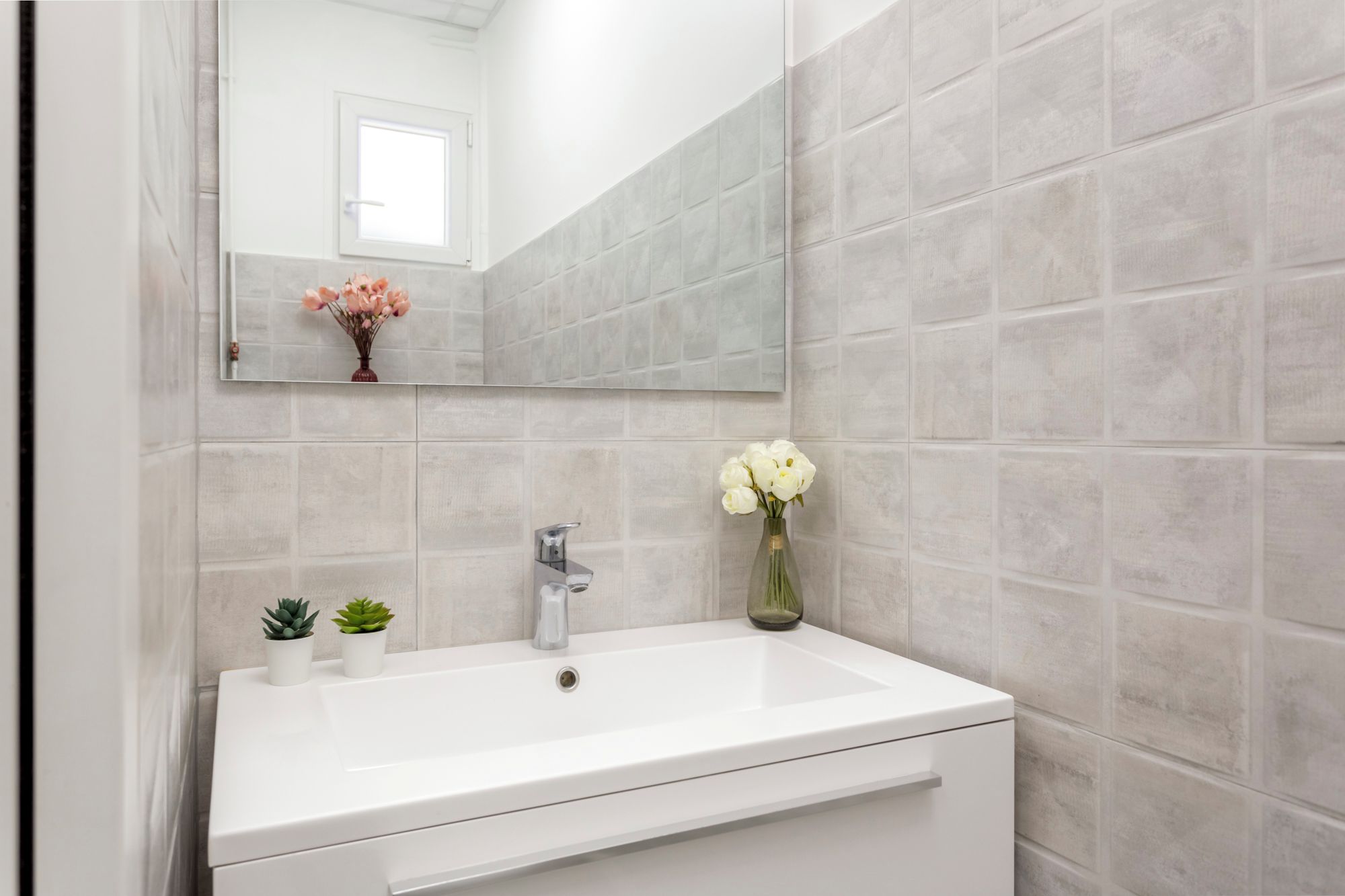 Une salle de bain avec un lavabo blanc, un miroir et un vase contenant des fleurs blanches.