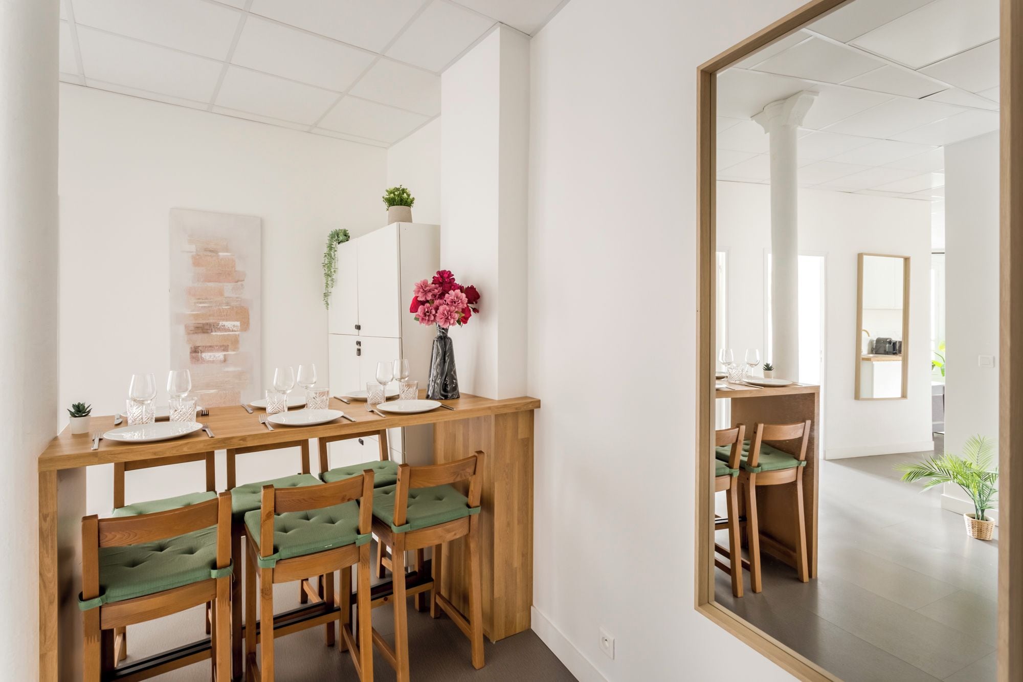 Een eethoek met een houten tafel voor zes personen, zes stoelen met groene kussens, een vaas met roze bloemen en een wit kastje met een plant.