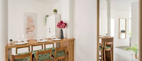 Un comedor con una mesa de madera dispuesta para seis comensales, seis sillas con cojines verdes, un jarrón con flores rosas y un armario blanco con una planta.