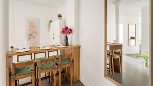 Un coin repas avec une table en bois dressée pour quatre personnes, entourée de chaises à coussins verts. Un vase rempli de fleurs roses est posé sur la table, et une plante en pot est accrochée au mur.