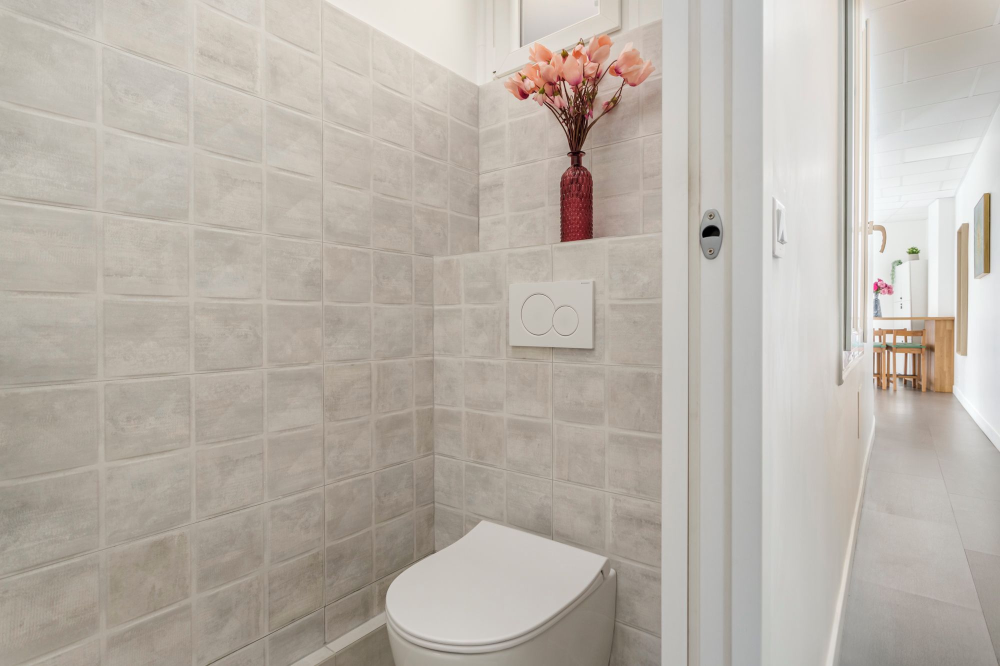 Bagno con piastrelle color crema, un water bianco, pulsante a muro e un vaso con fiori appeso alla parete.