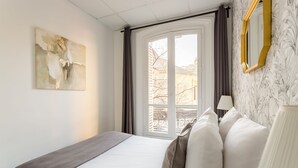 Une chambre d’hôtel avec un lit, une fenêtre avec des rideaux, une lampe et un mur orné d’un miroir encadré.