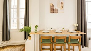 Un coin repas avec une table en bois dressée pour quatre personnes, deux chaises dotées de coussins verts, une plante en pot et une œuvre d’art encadrée accrochée au mur.