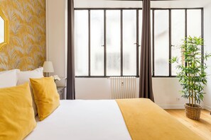Ein Hotelzimmer mit einem Bett, einem Fenster mit Jalousien, einem Heizkörper, einem Nachttisch mit Lampe und einer Zimmerpflanze.