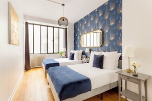 Une chambre d’hôtel avec deux lits, une grande fenêtre avec des rideaux, un radiateur, une table de chevet avec une lampe et un miroir au mur.