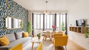 Een moderne woonkamer met een bank, gele fauteuils, een eettafel met stoelen, een televisie en een groot raam met gordijnen.