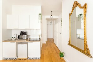 A modern kitchen with white cabinets, a microwave, and a coffee maker. There is a hallway with a wooden floor and a large ornate mirror on the wall. - Merveil - Family Suite - Louvre - Sentier I (Paris)