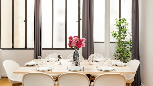 Een eetgedeelte met een lange tafel gedekt voor vier personen, witte stoelen, een vaas met roze bloemen en een plant in pot.