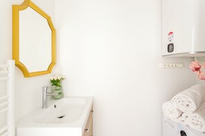 Ein Badezimmer mit einem weißen Waschbecken, einem spiegel mit gelbem Rahmen, einer grünen Vase mit weißen Blumen und einem Stapel weißer Handtücher.