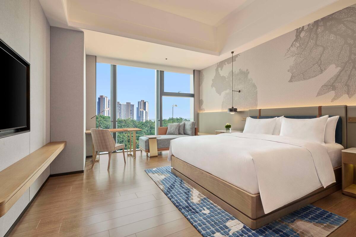 A modern hotel room with a large bed, a desk, a chair, a TV, and a city view.