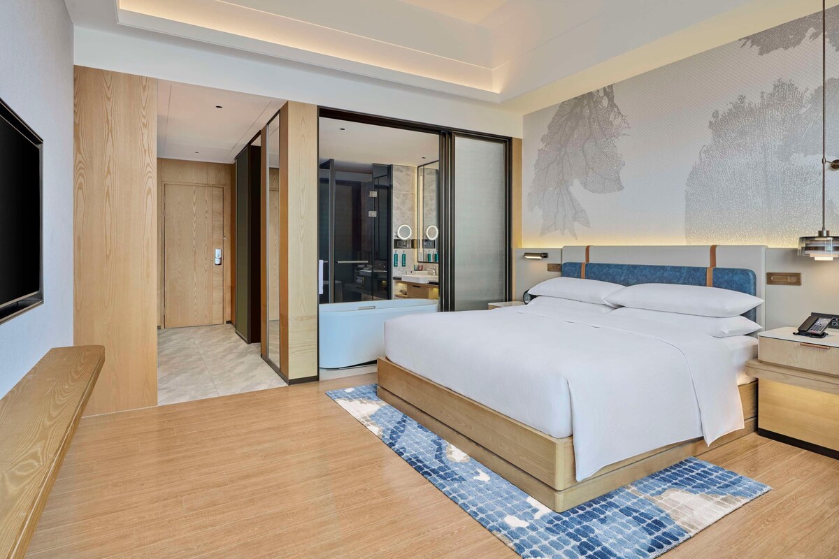 A modern hotel room with a large bed, a wooden bench, a desk, and a bathroom visible through a sliding door.