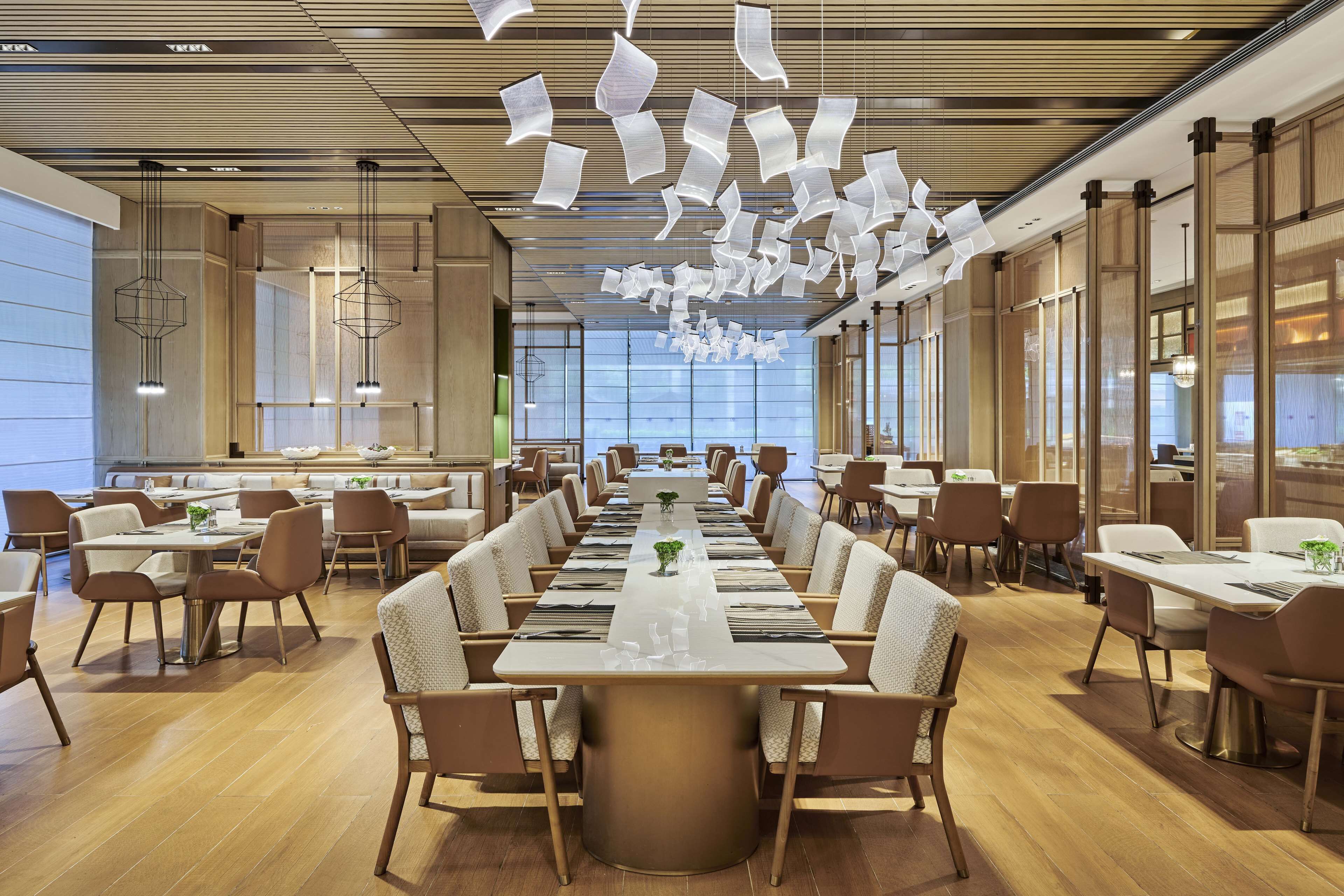 A spacious dining area with long tables and chairs, a central decorative light fixture, and large windows allowing natural light.