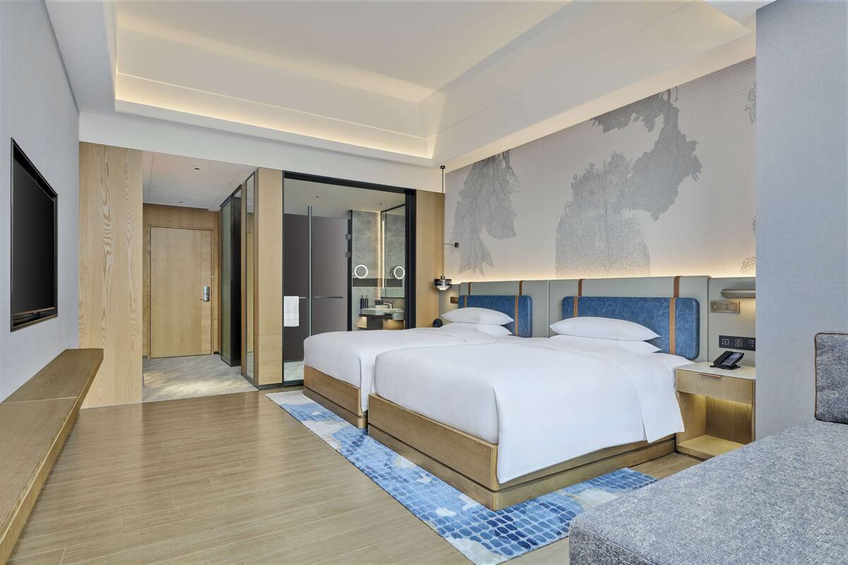A modern hotel room with two beds, a flat-screen TV, a large mirror, and a wall-mounted artwork.