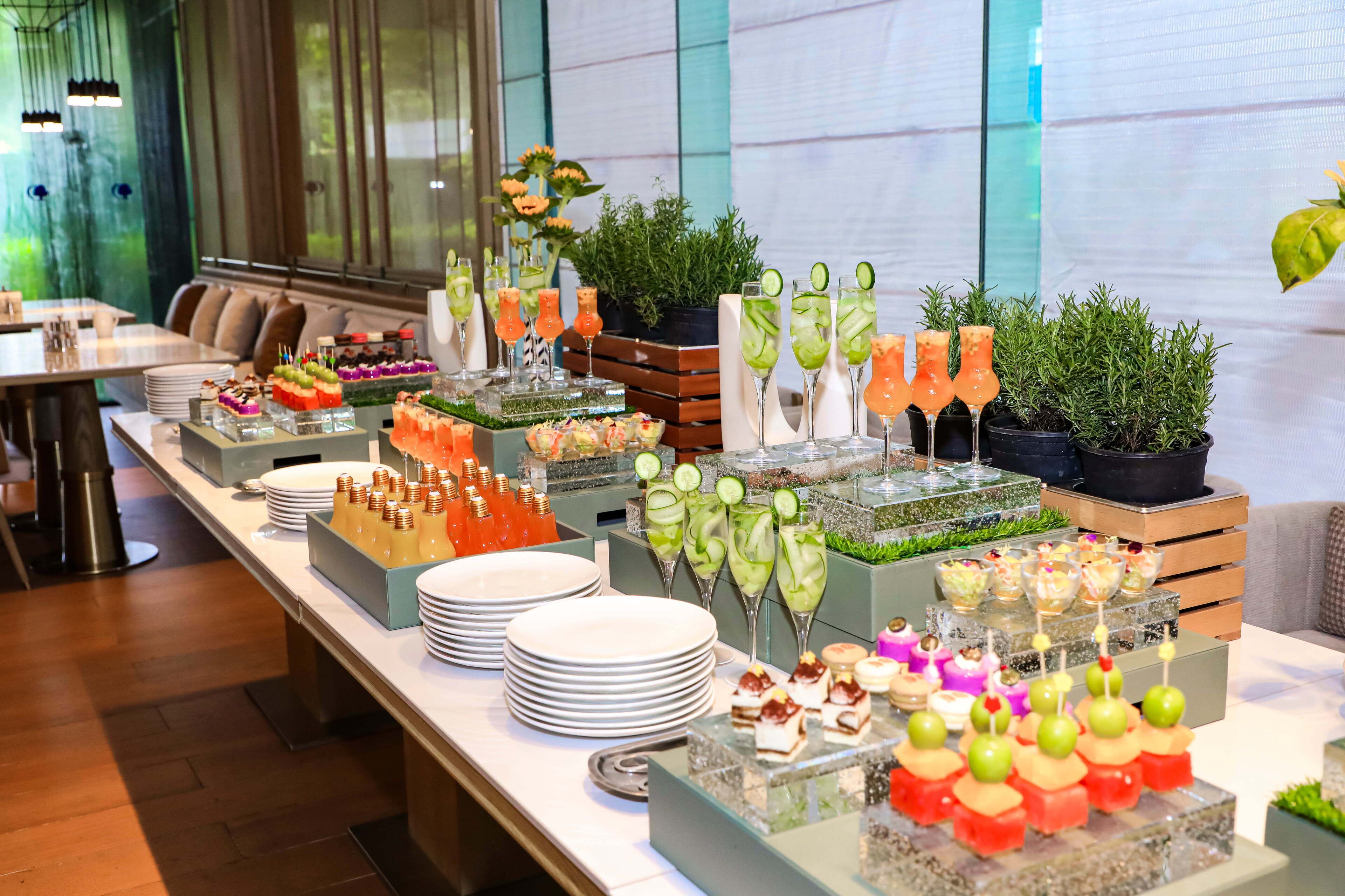 Un bufé con diversos alimentos y bebidas, entre ellos brochetas de fruta, ensaladas y postres, dispuesto en una larga mesa en una sala bien iluminada con ventanas grandes.