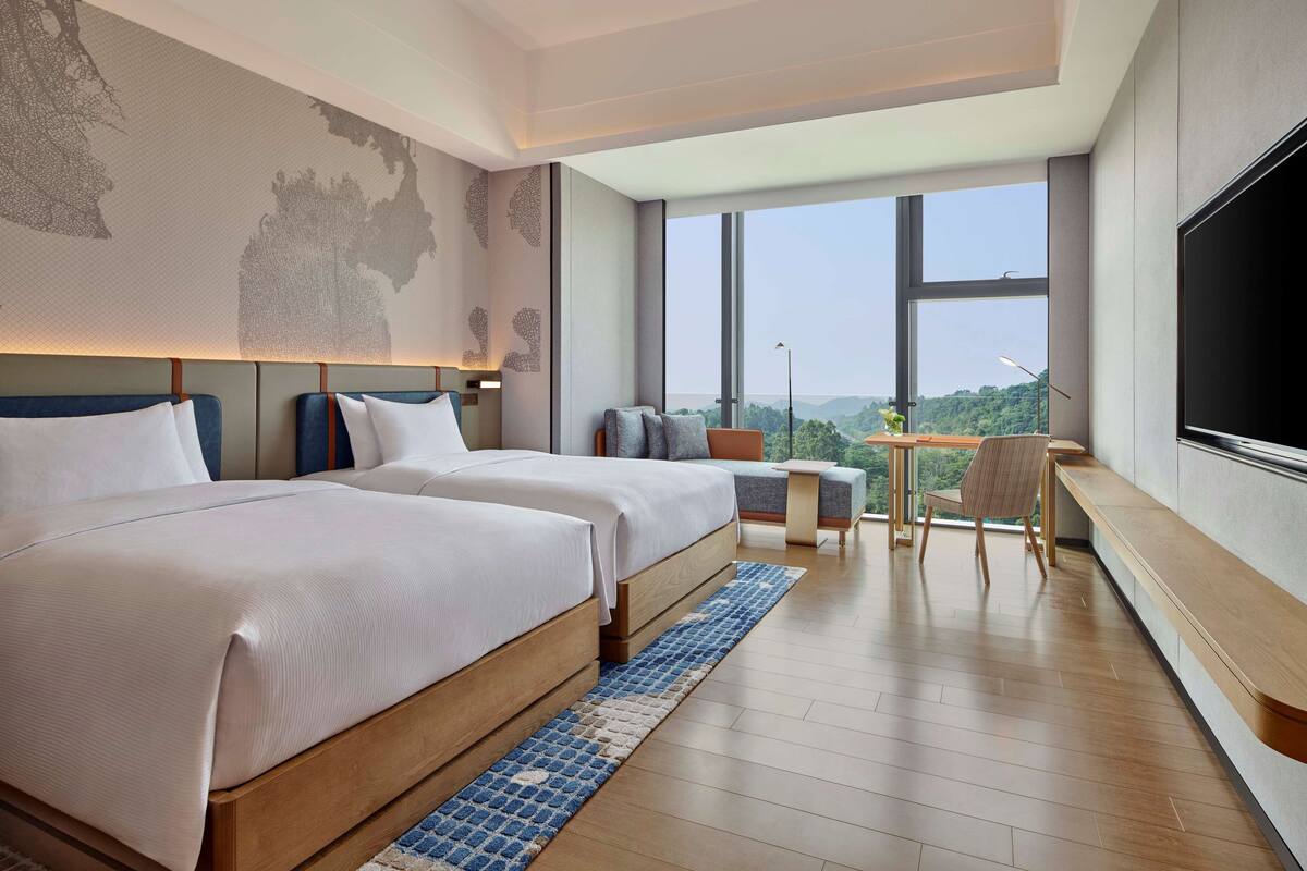 A modern hotel room with two beds, a desk, and a large window offering a city view.