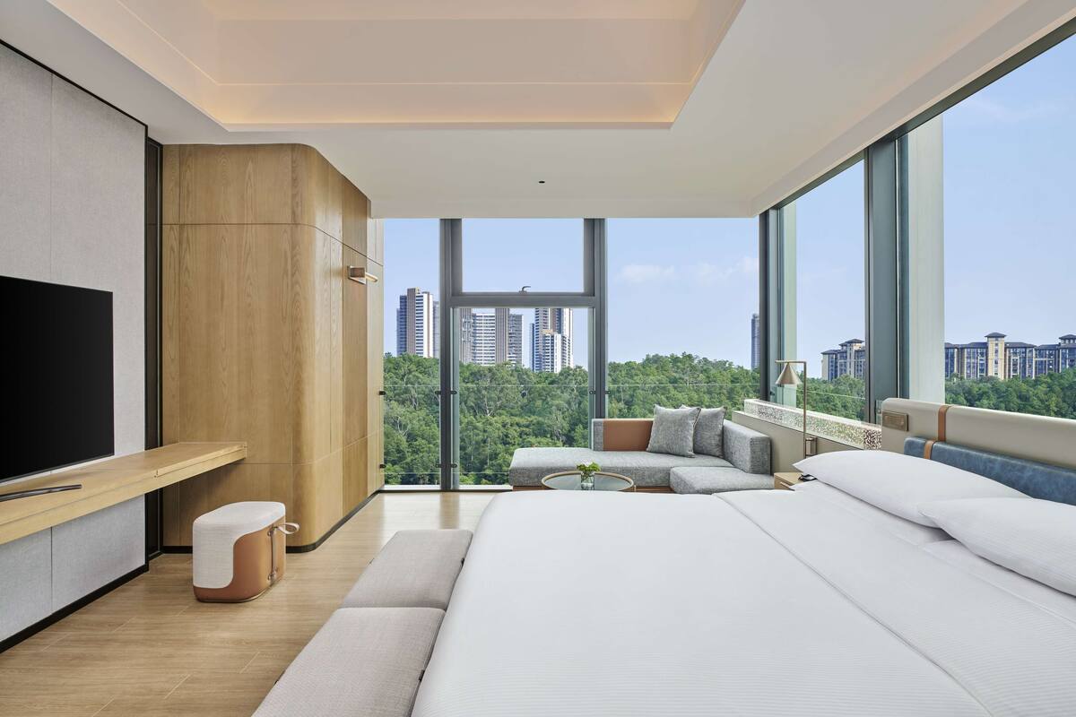 A modern hotel room with a large bed, a TV, a wooden wardrobe, and a view of the cityscape.